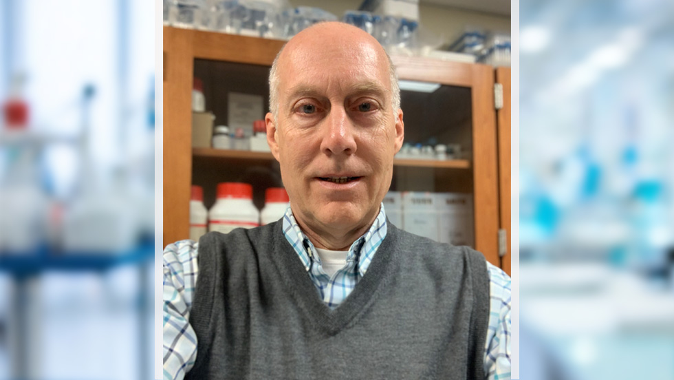 A man, smiling, stands in front of a cabinet with chemistry supplies