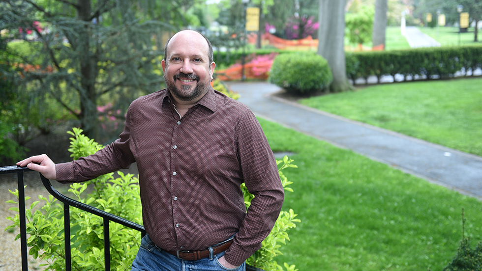 A man in a brown shirt, smiling, standing outside next to an iron stair rail, trees and lawn in background