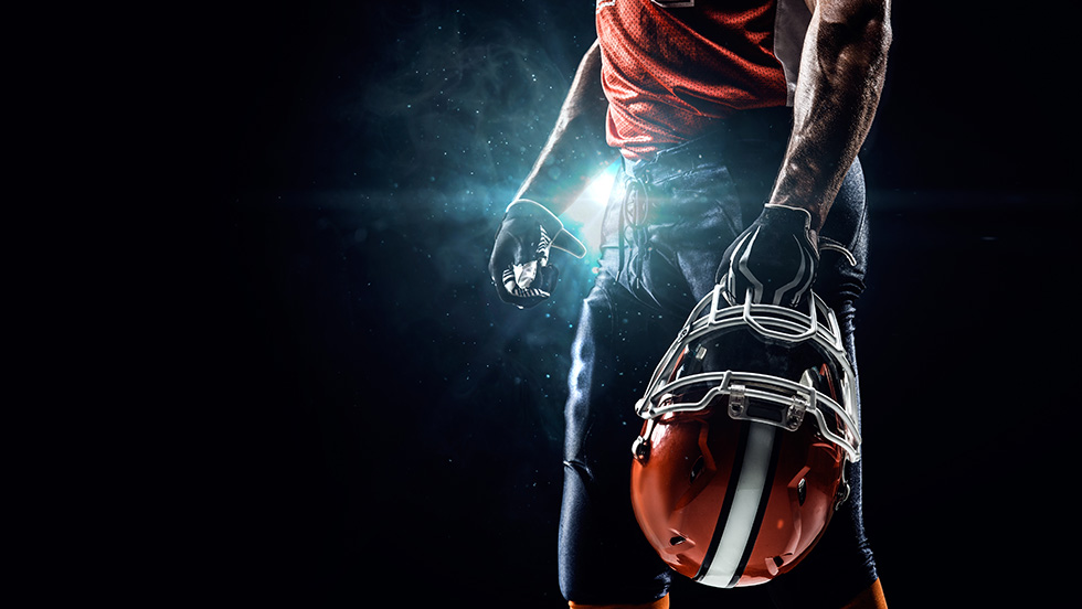 An evocative photo of a football player holding his helmet at his side. The player is seen only from his midsection to his thighs. It is night, so the background is black except for light from a single floodlight behind the player.