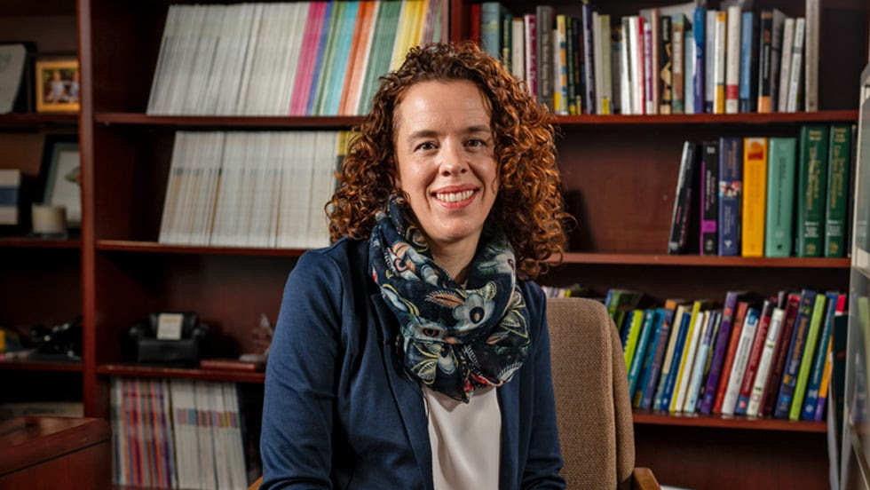 Woman with reddish hair sitting in front of bookshelf.