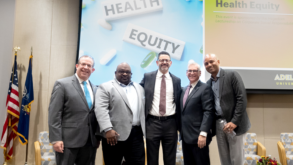 Five gentlemen in suits stand before "Health Equity" signage, with two flags and chairs visible behind them.