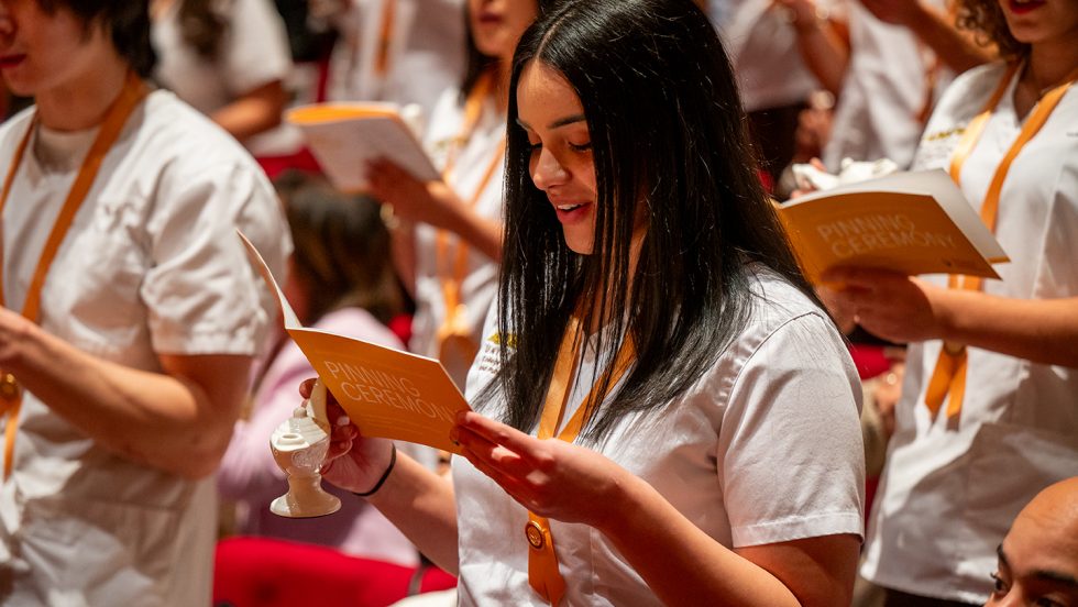 Students wearing white scrubs and reading a yellow book during pinning ceremony.