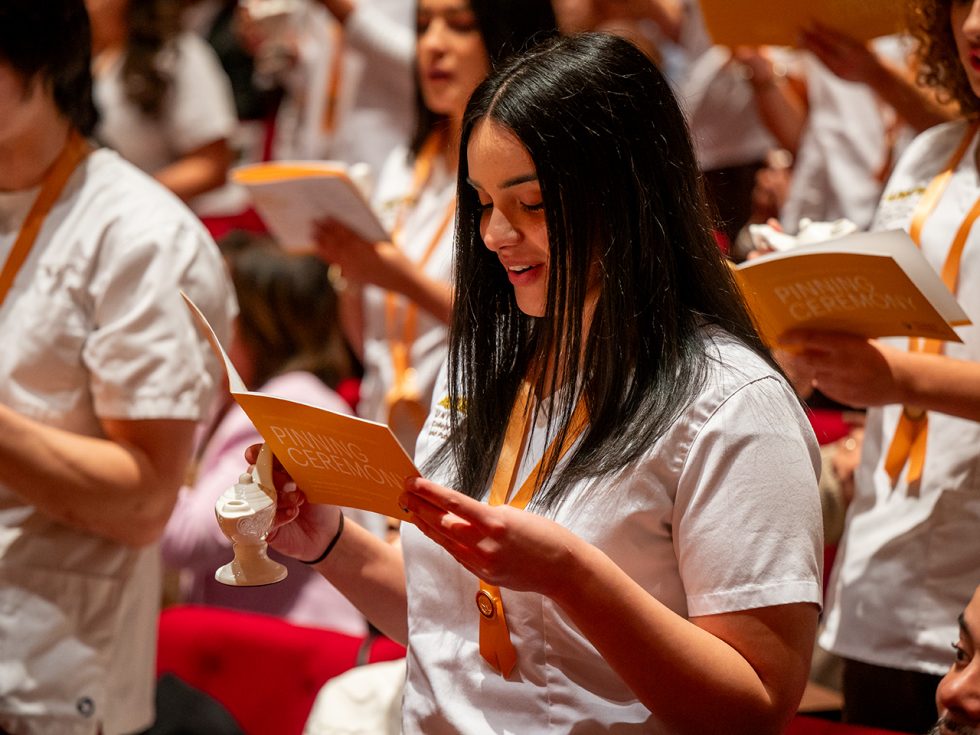 Students wearing white scrubs and reading a yellow book during pinning ceremony.