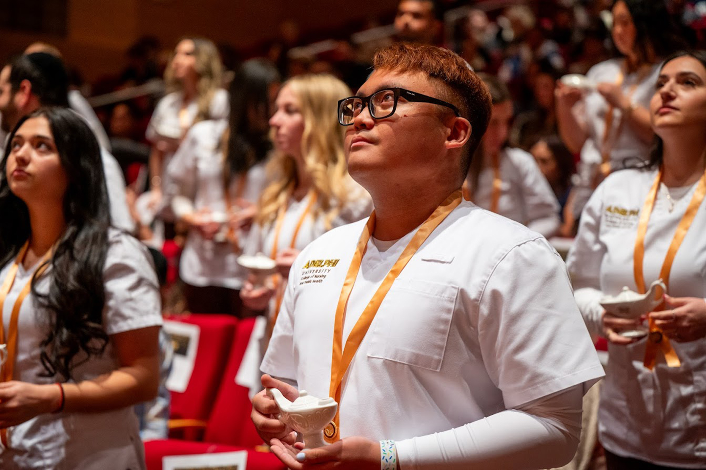 A crowd of students wearing nursing coats in an auditorium, each holding a small white lamp.