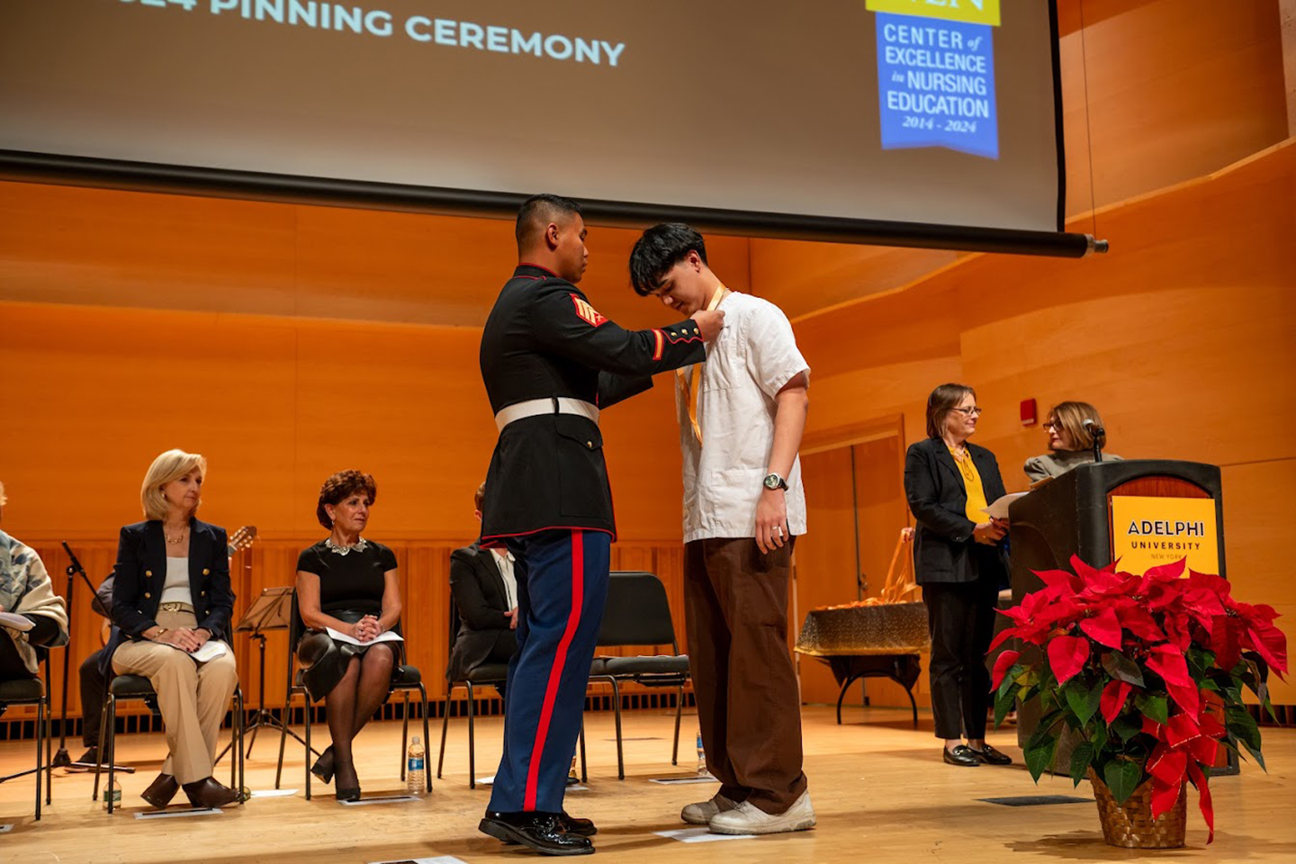 A man in a military uniform places a medal around the neck of a student in a nursing coat on a stage.