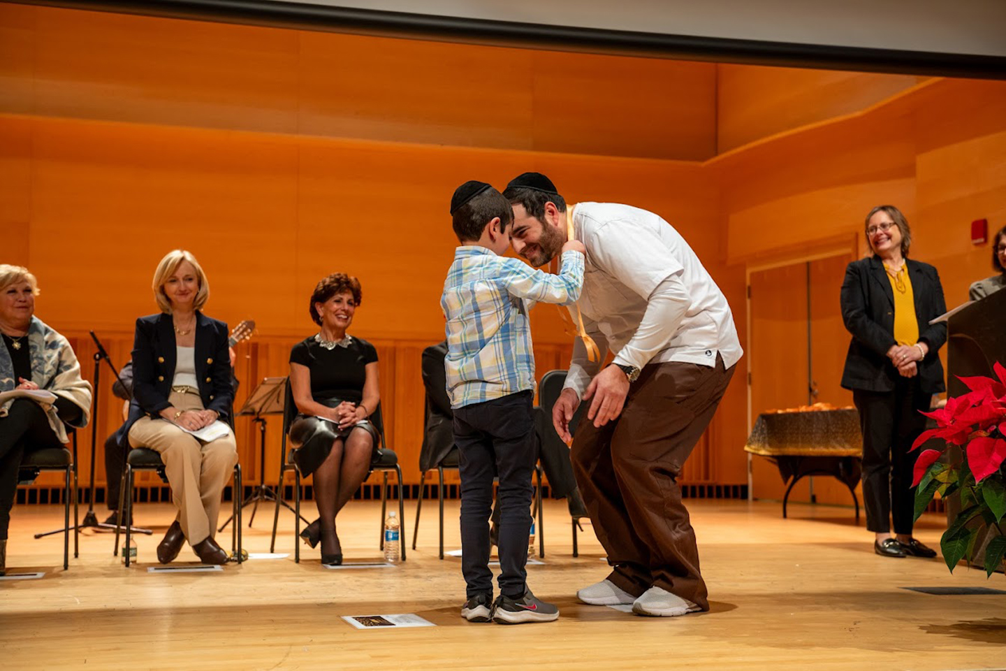 A child places a medal around the neck of a hunched-over man in a nursing coat on a stage.