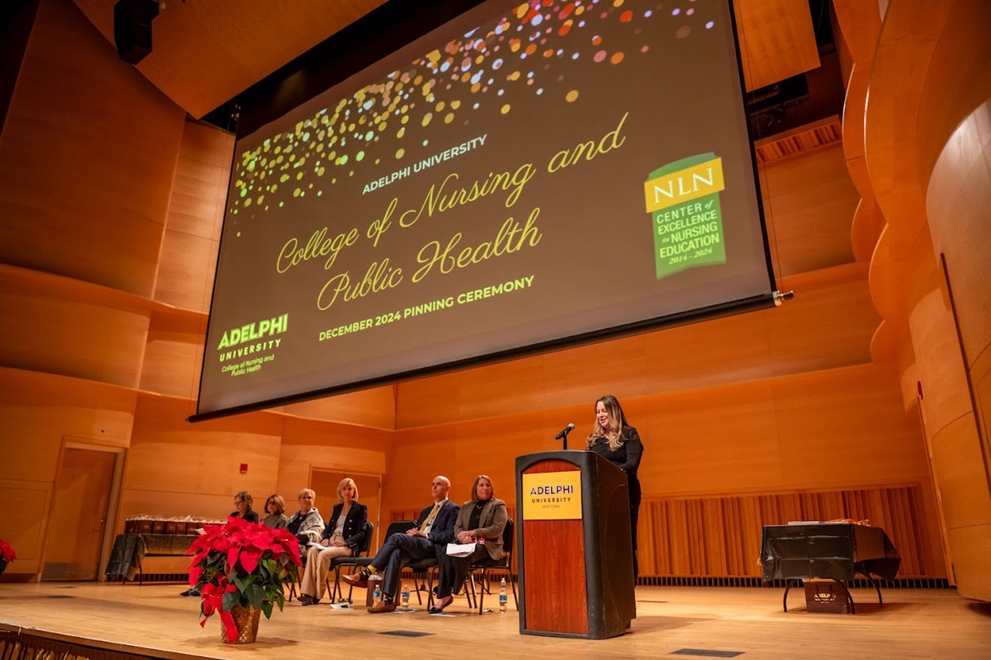 A lineup of people sitting on a stage listen to a woman at a podium. A screen reading “College of Nursing and Public Health” hangs above the stage.