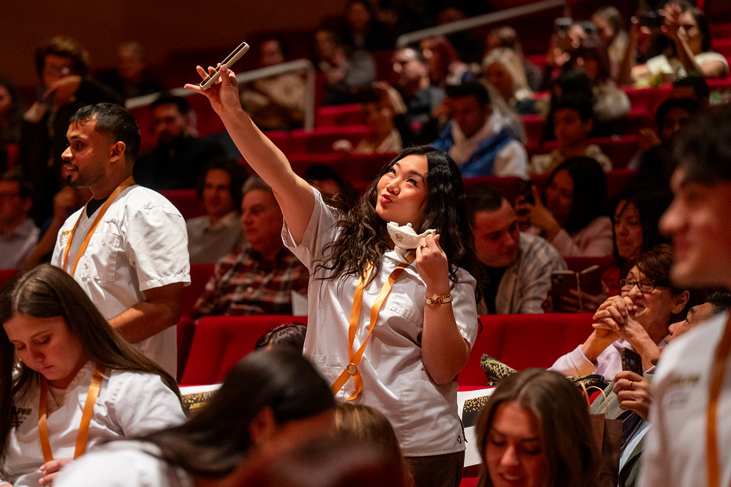 A woman in a nursing coat standing in front of her seat in an auditorium takes a selfie of herself and a small white lamp.