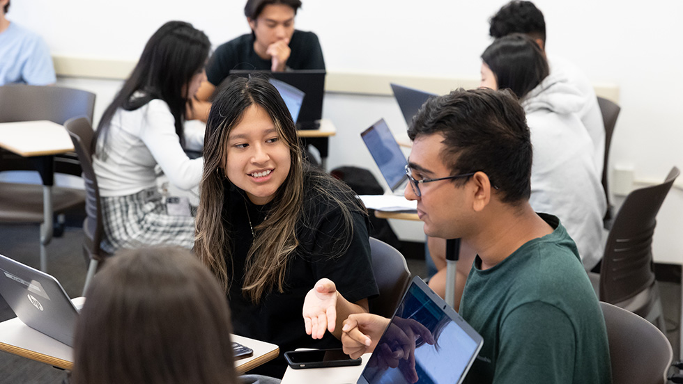 Three students sit at a table with their computers, engaging in conversation, with another group of four students doing similar work in the background.