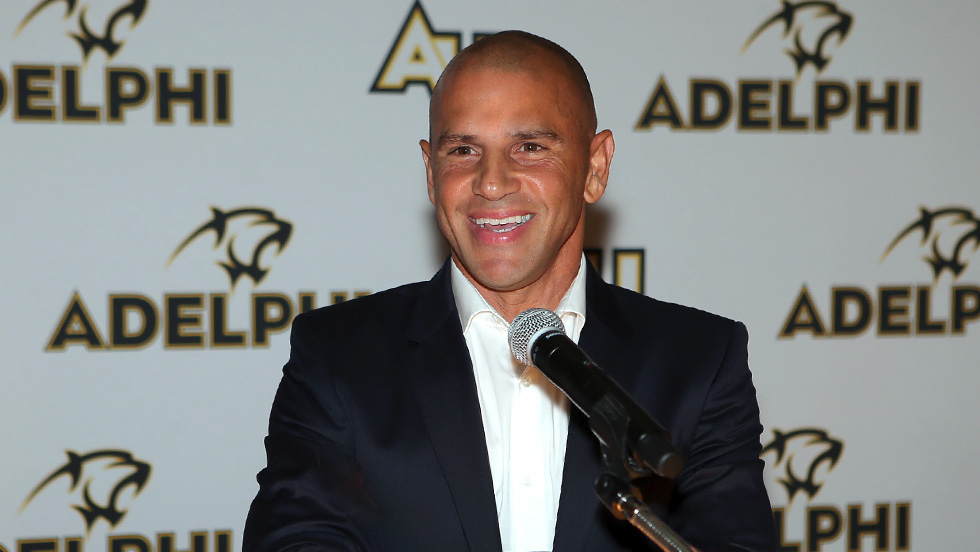 Chris Armas stands at the microphone in front of an Adelphi University backdrop.