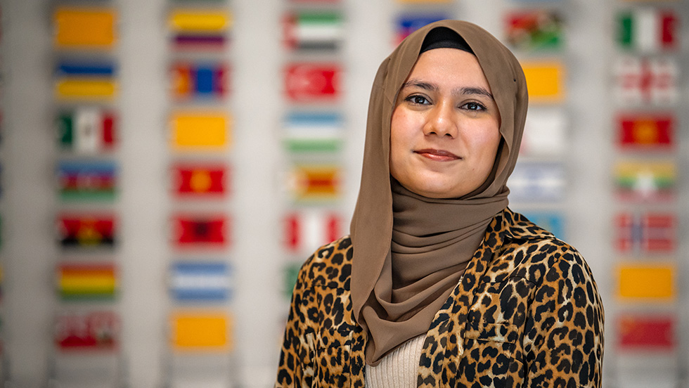 Muneera Chowdhury, wearing a hijab, the headscarf worn by Muslim women, stands in front of a wall with the flags of all international students at Adelphi.