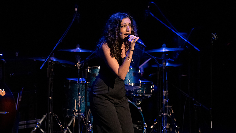 Veronica Mansour, microphone in hand, sings on stage. A drum set is behind her.