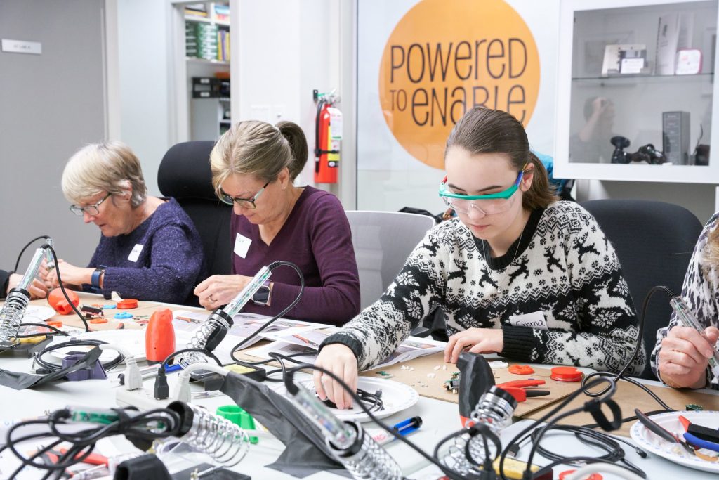 Three women of various ages sitting at a table working with tools. 