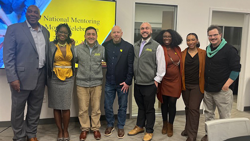 A group of eight mentoring leaders gather together in a row in front of a large screen that reads, National Mentoring Night Celebration. They are happy, proud and smiling.