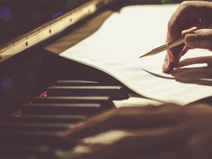 Hands and a sheet of paper on a piano keyboard; right hand holds a pencil.