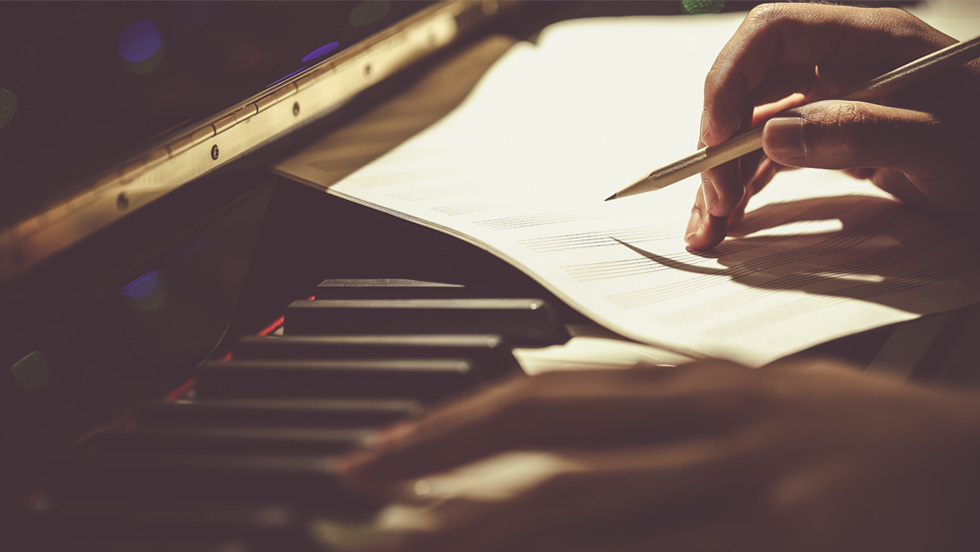 Hands and a sheet of paper on a piano keyboard; right hand holds a pencil.