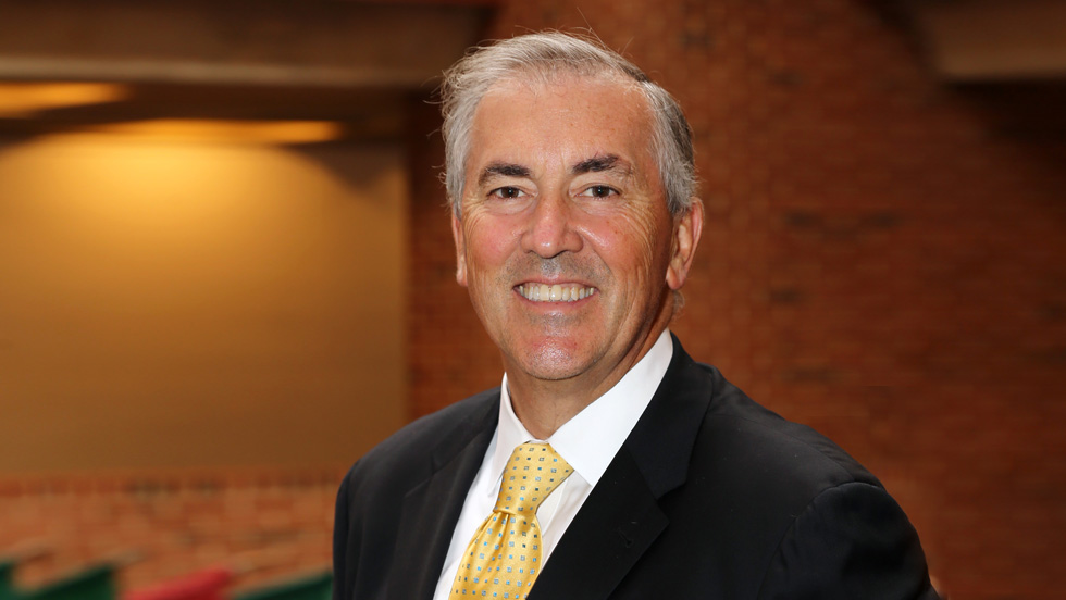 A man wearing a suit and tie stands against a neutral wall, representing a professional and sophisticated demeanor.