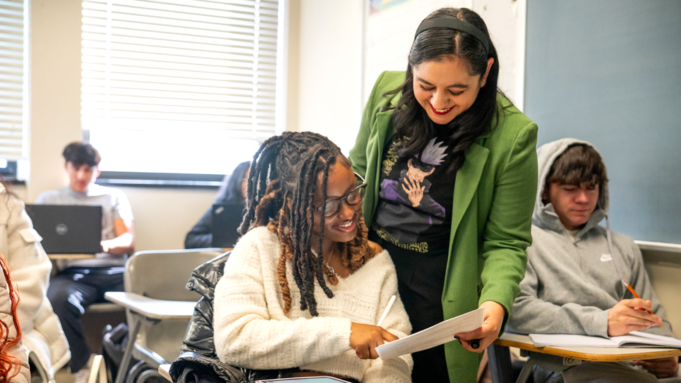 Woman teacher standing in classroom of students, showing a paper to a student sitting at a desk.